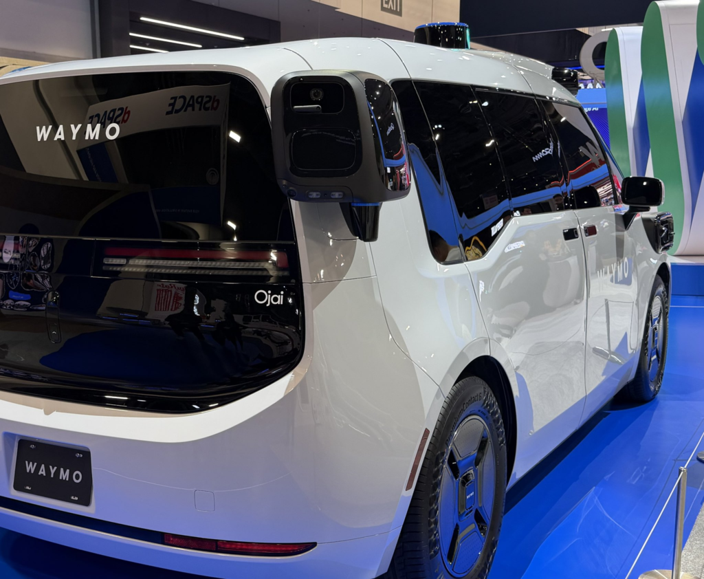 A white Waymo 6th-generation vehicle, Ojai, stationed in a Waymo parking lot.